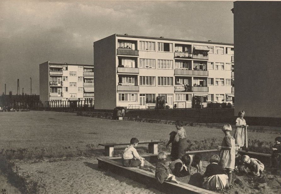 Schwarz-Weiß-Fotografie mit spielenden Kindern in einem Sandkasten im Vordergrund und Mehrfamilien-Siedlungshäusern im Hintergrund
