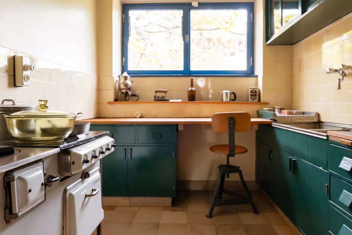 Foto mit Blick in eine kleine, funktionale Küche mit Herd, Fensterfront und Stuhl