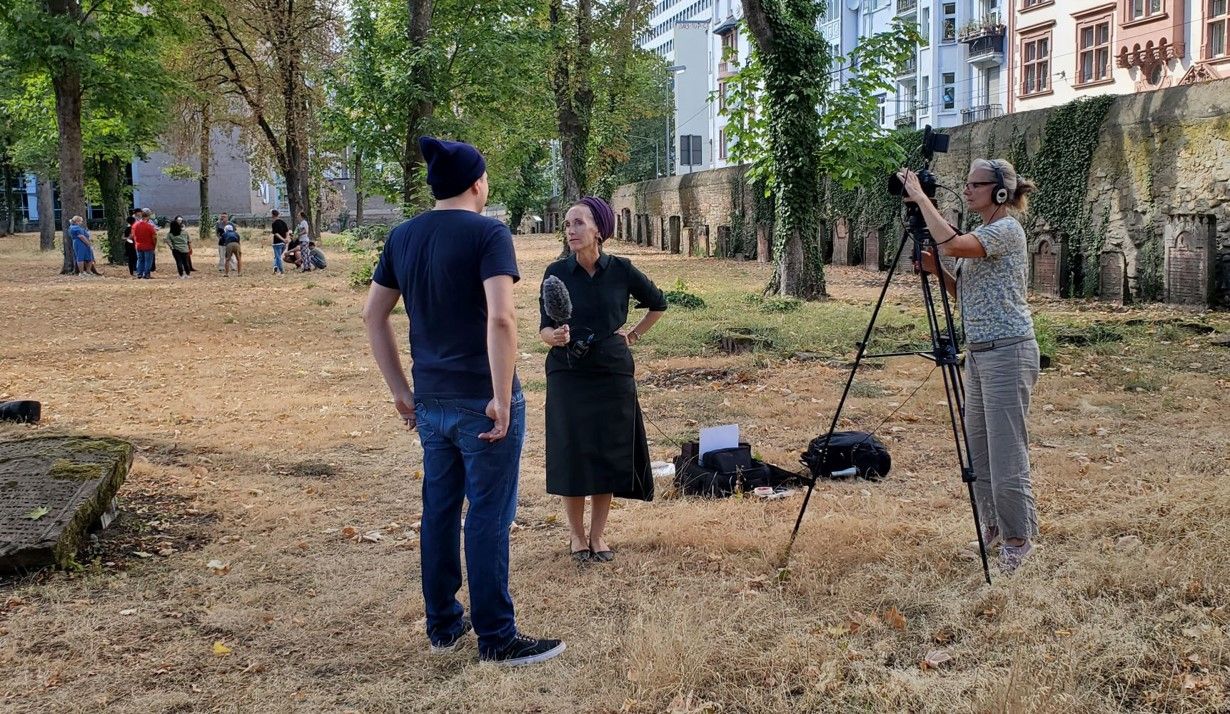 Foto einer Interviewsituation, ein Mann steht mit dem Rücken zum Betrachter auf einem Friedhof und wird von einer Frau mit Mikro interviewt, eine weitere Frau rechts im Bild filmt